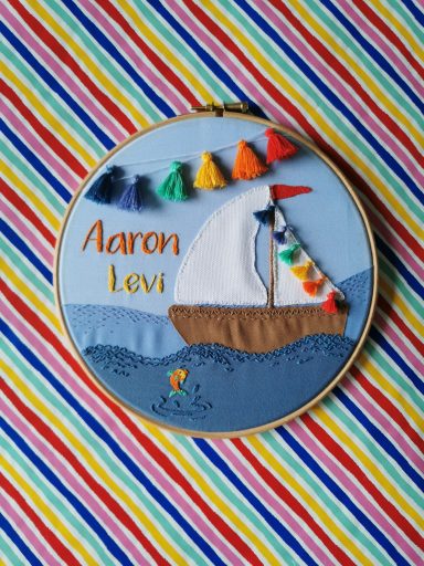 Stickbild zur Geburt eines Kindes mit dem Namen Aaron Levi. Zu sehen ist ein kleiner Fisch und ein Boot mit bunter Girlande im Wasser.